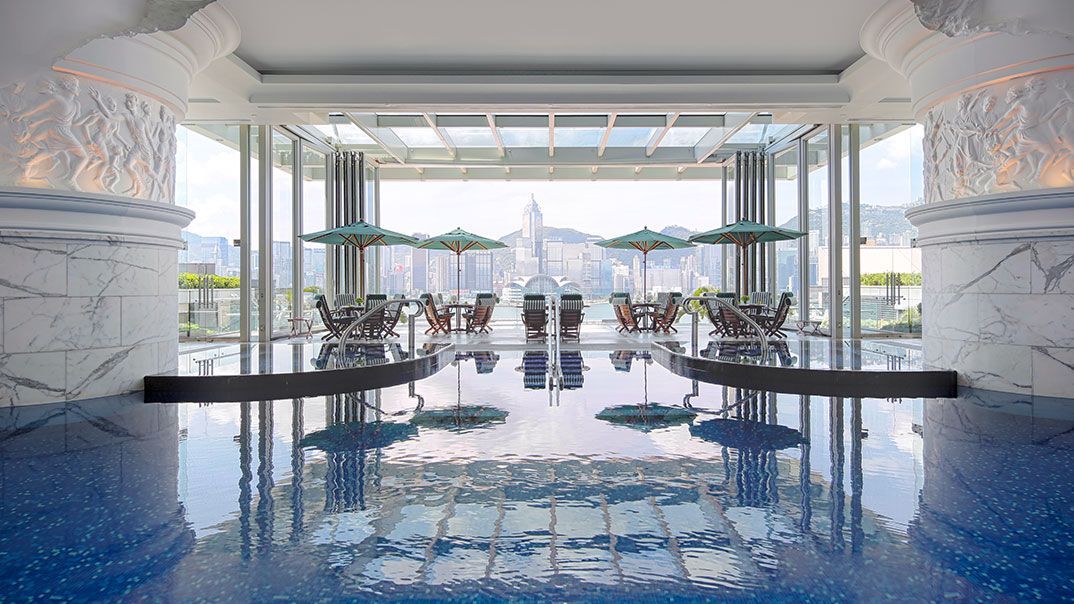 A luxurious pool reflects an open-air dining area with city views. Blue water, marble columns, tables, and umbrellas. The Langham, Hong Kong