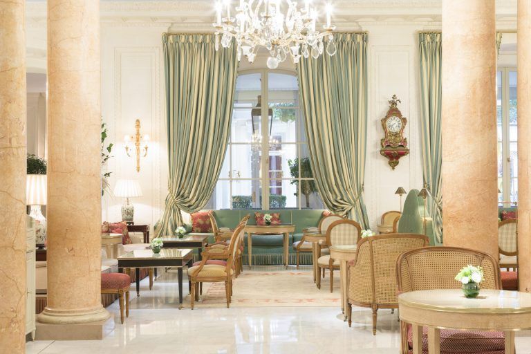 An elegant hotel lounge featuring classic columns, green draped windows, chandeliers, and arranged seating. Le Bristol Paris