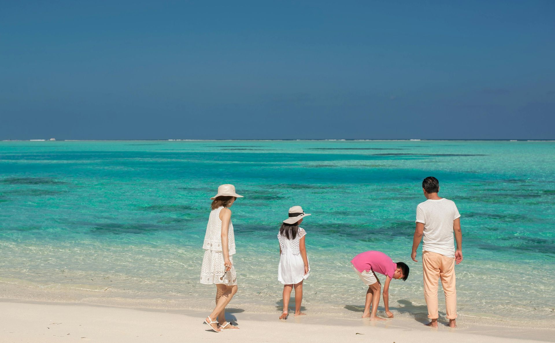 A family stands on a white sandy beach by crystal clear turquoise water under a bright blue sky.