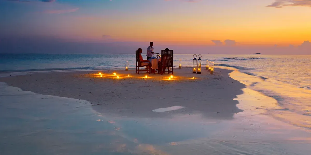 Romantic dinner setting on a beach at sunset, with candles, table, and two people.