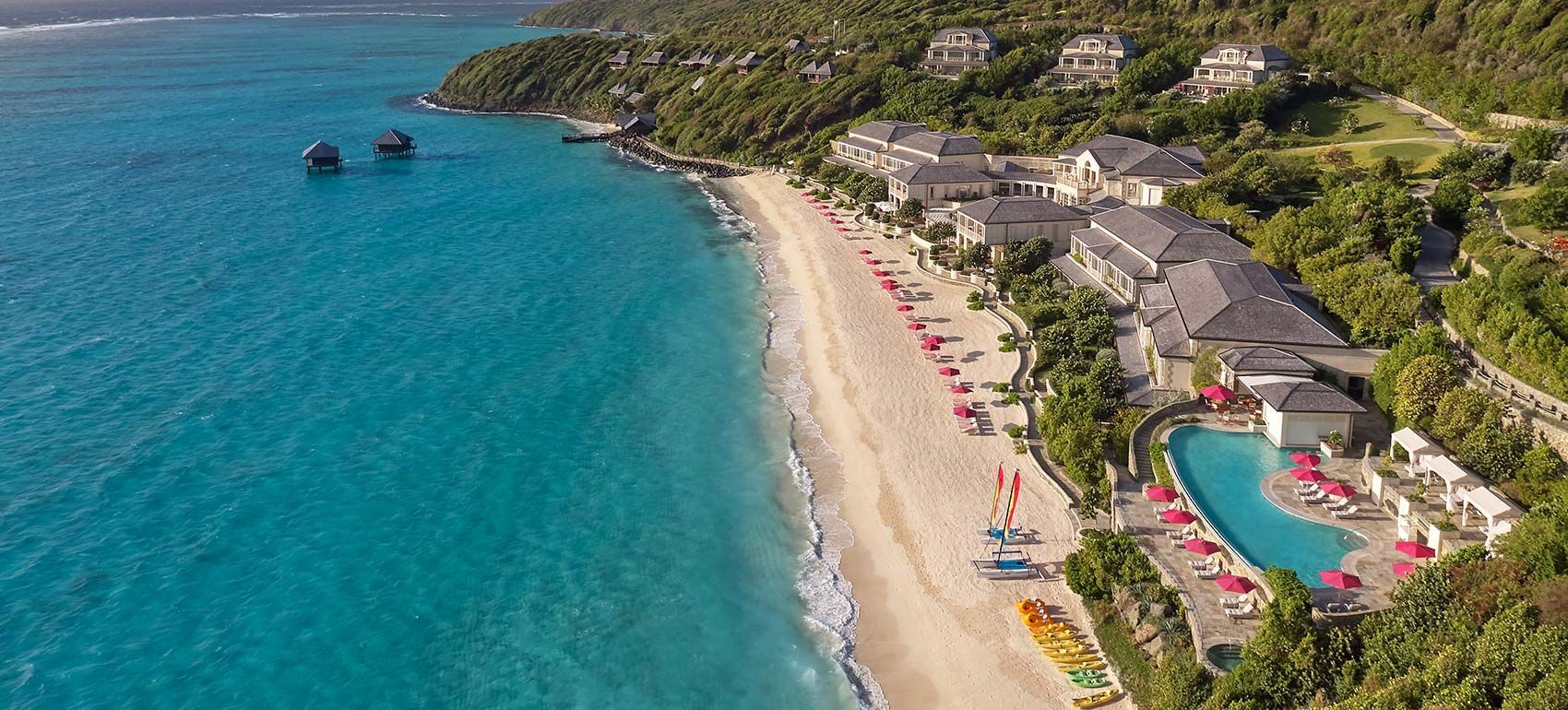 Aerial view of a beach with turquoise water, white sand, and a resort. Lush green trees and buildings surround the coast. Mandarin Oriental, Canouan