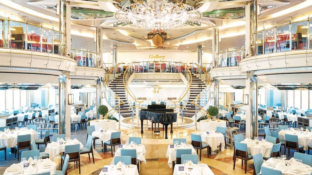 Elegant cruise ship dining room with tables, grand piano, and dual staircases.