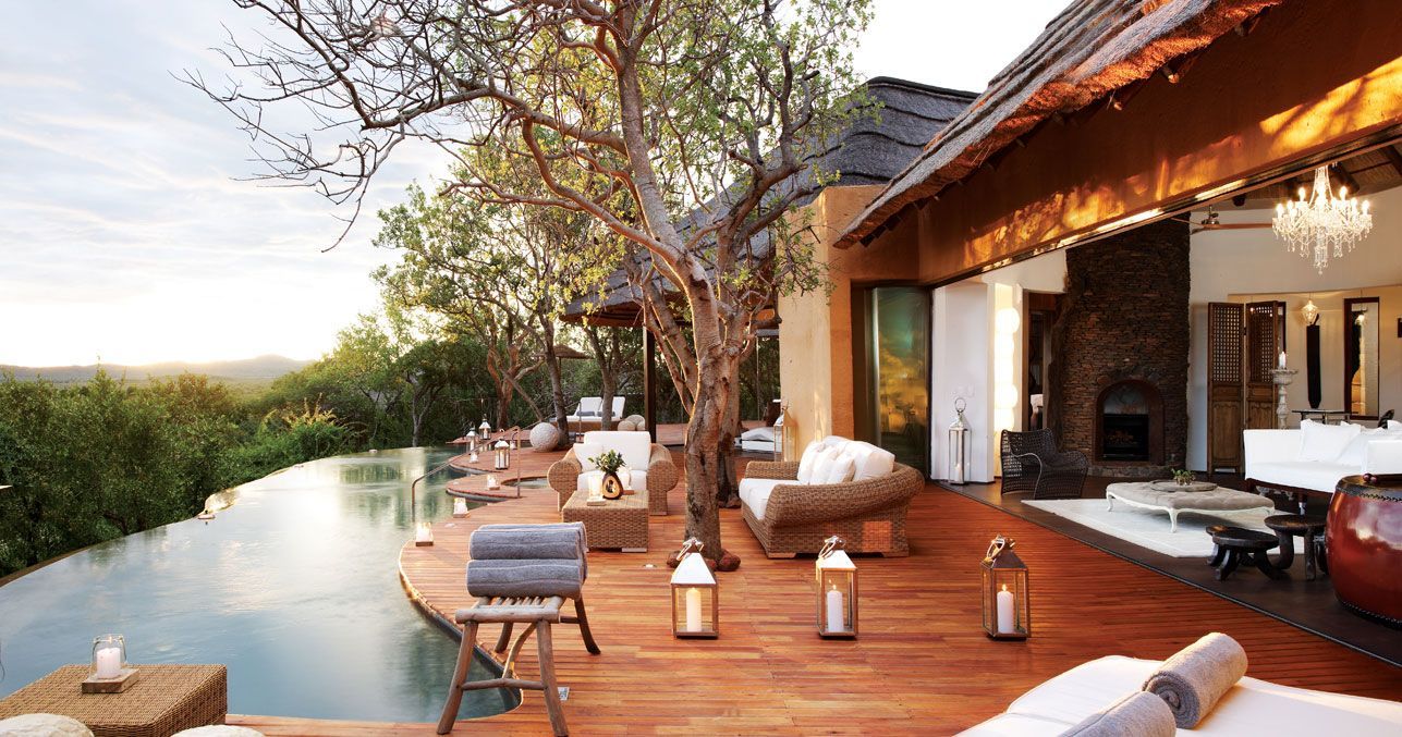 Wooden deck with infinity pool, luxury lodge with open doors, overlooking a lush landscape.