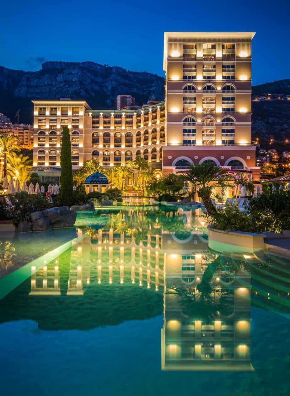 Luxury hotel at dusk, reflected in pool. Lights illuminate building, mountains in background.