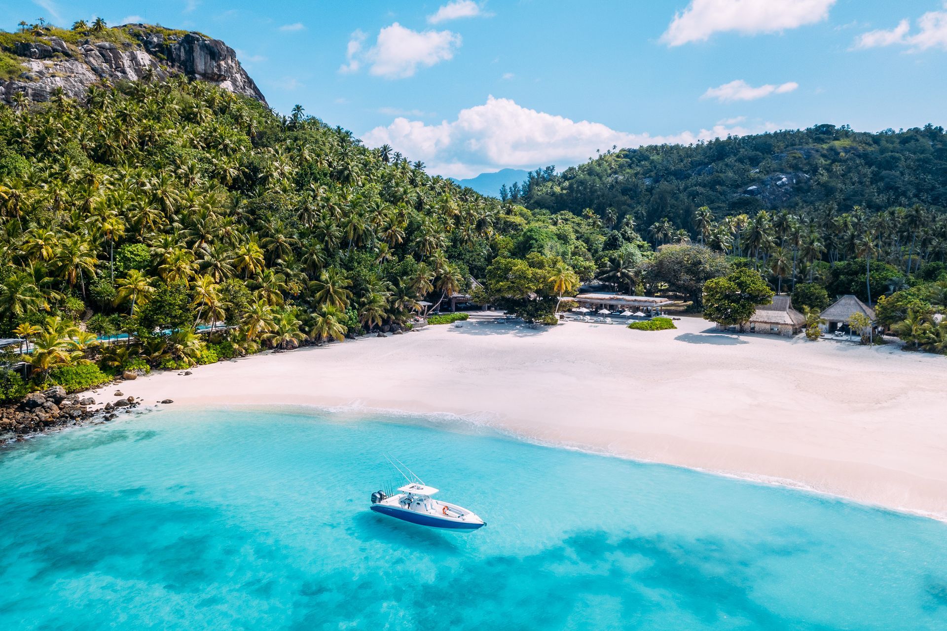 A boat anchored in bright turquoise water off a white sand beach bordered by a lush tropical forest and mountains. north Island