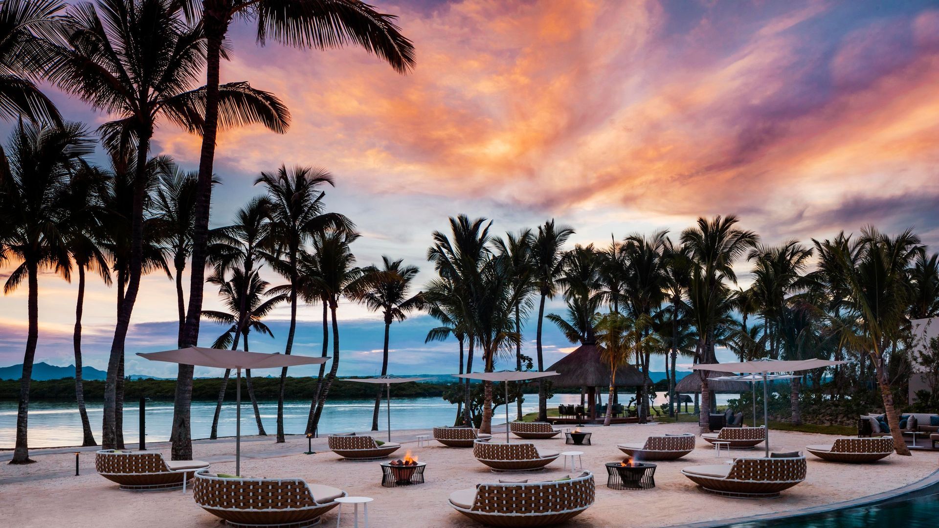 Luxury beach resort with palm trees, sun loungers, and umbrellas at sunset with a pink and orange sky. One&Only Le Saint Géran