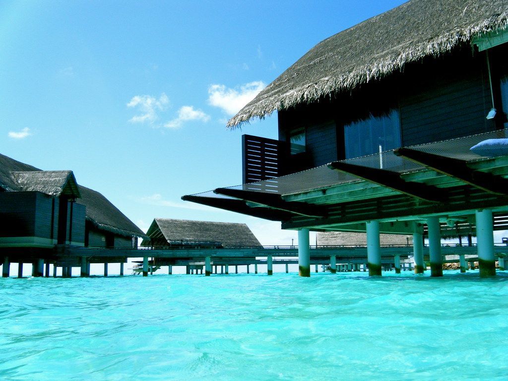 Overwater bungalows with thatched roofs over turquoise water under a bright blue sky. One&Only Reethi Rah
