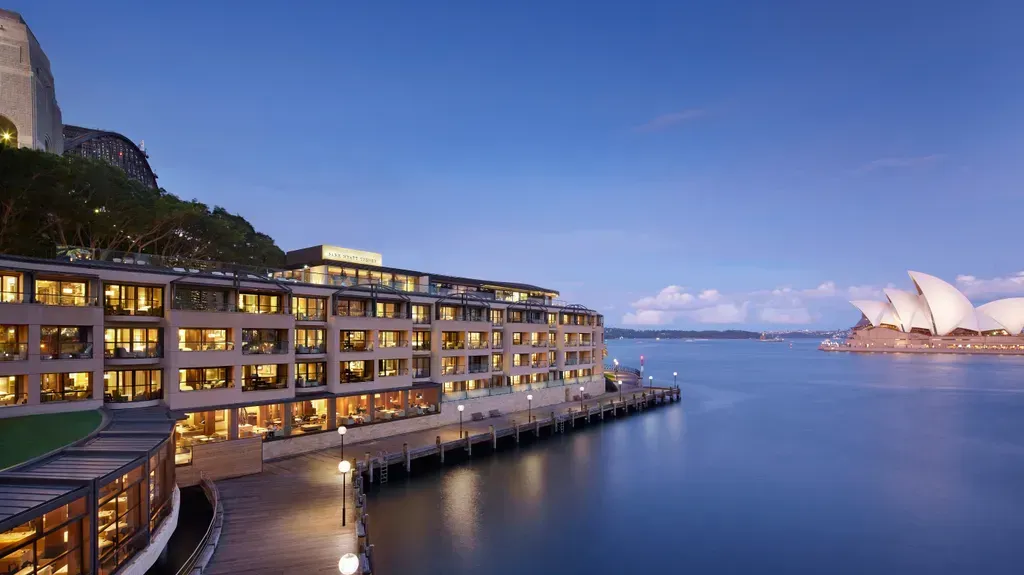 Hotel on waterfront, Sydney Opera House in the background. Calm blue water, dusk. 