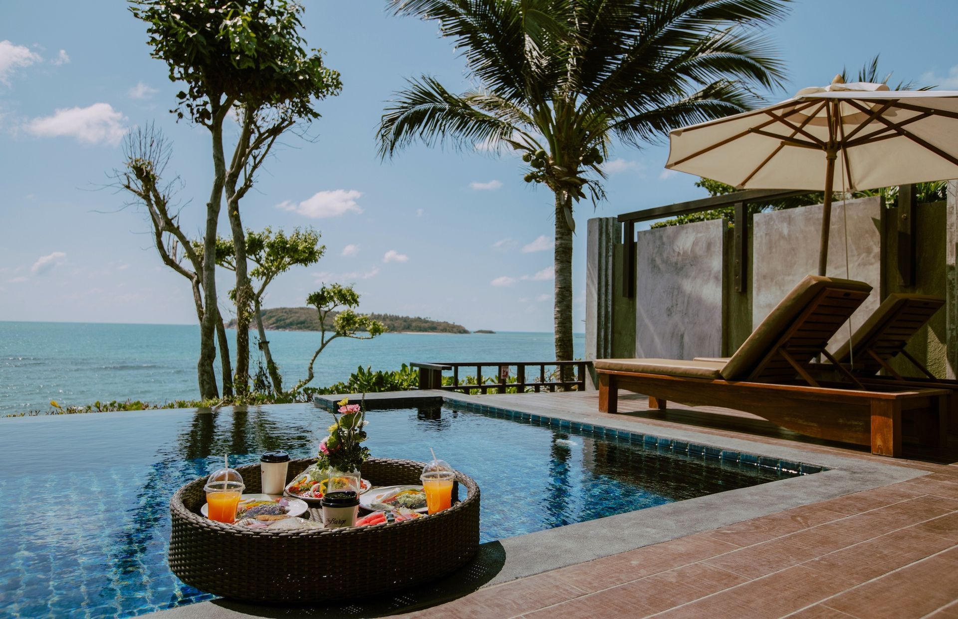 Poolside breakfast tray floating in a pool, ocean view, two lounge chairs, and an umbrella.