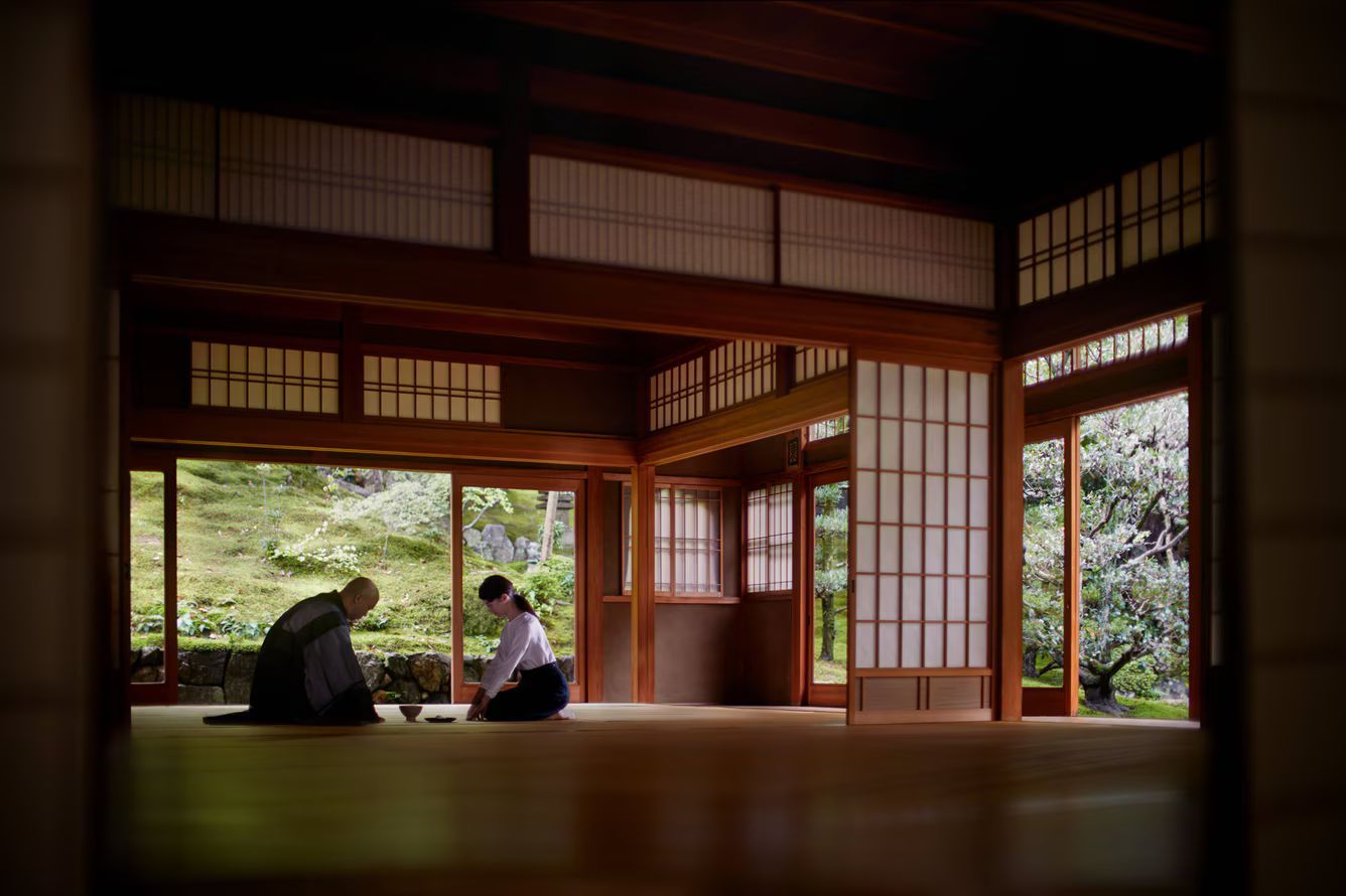 Two people sit on tatami mats in a traditional Japanese room, facing each other with an open garden view in the background.