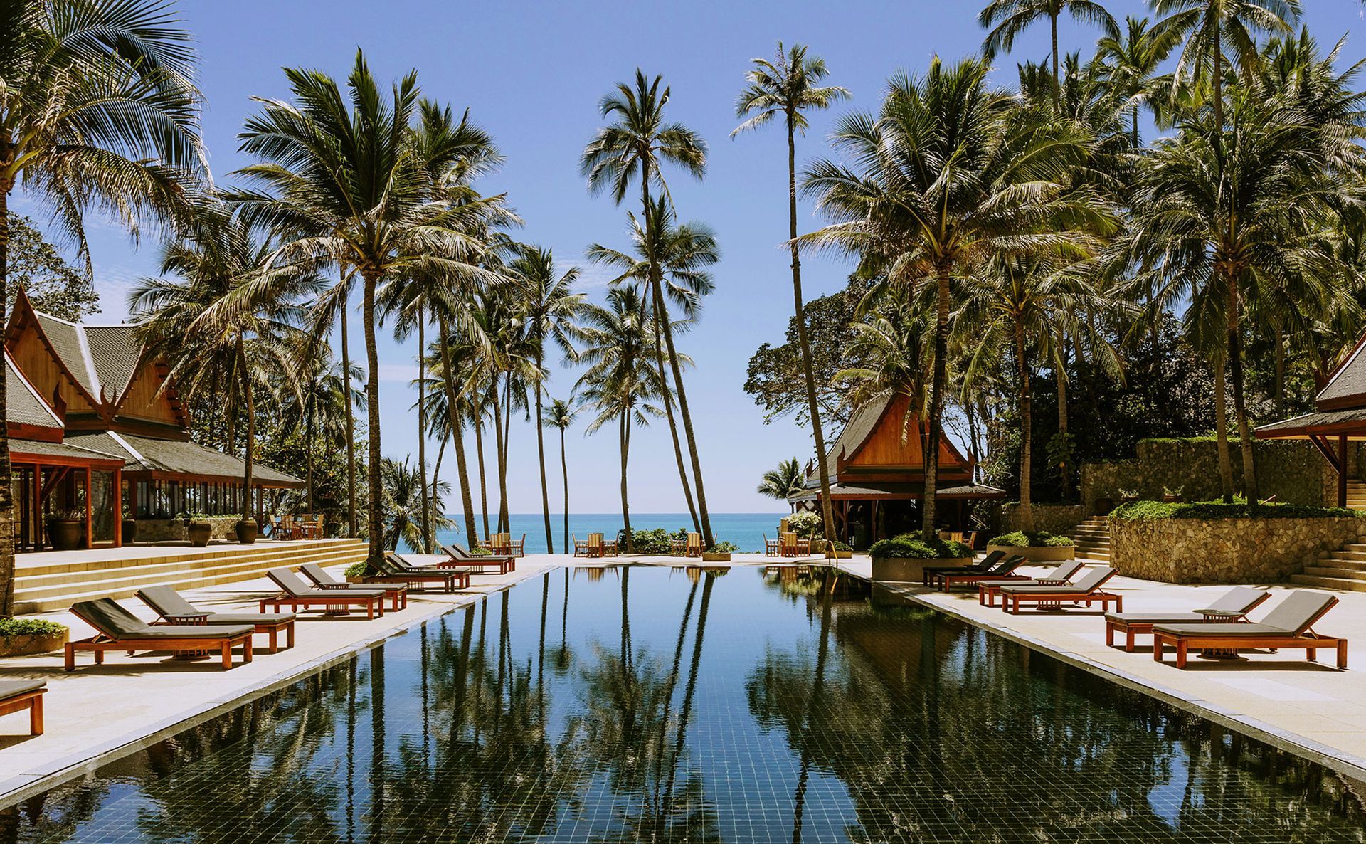 Pool lined with lounge chairs, palm trees, ocean view. Sunny resort setting.