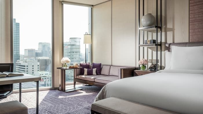 Modern hotel room with large windows, city view, bed, sofa, and desk
