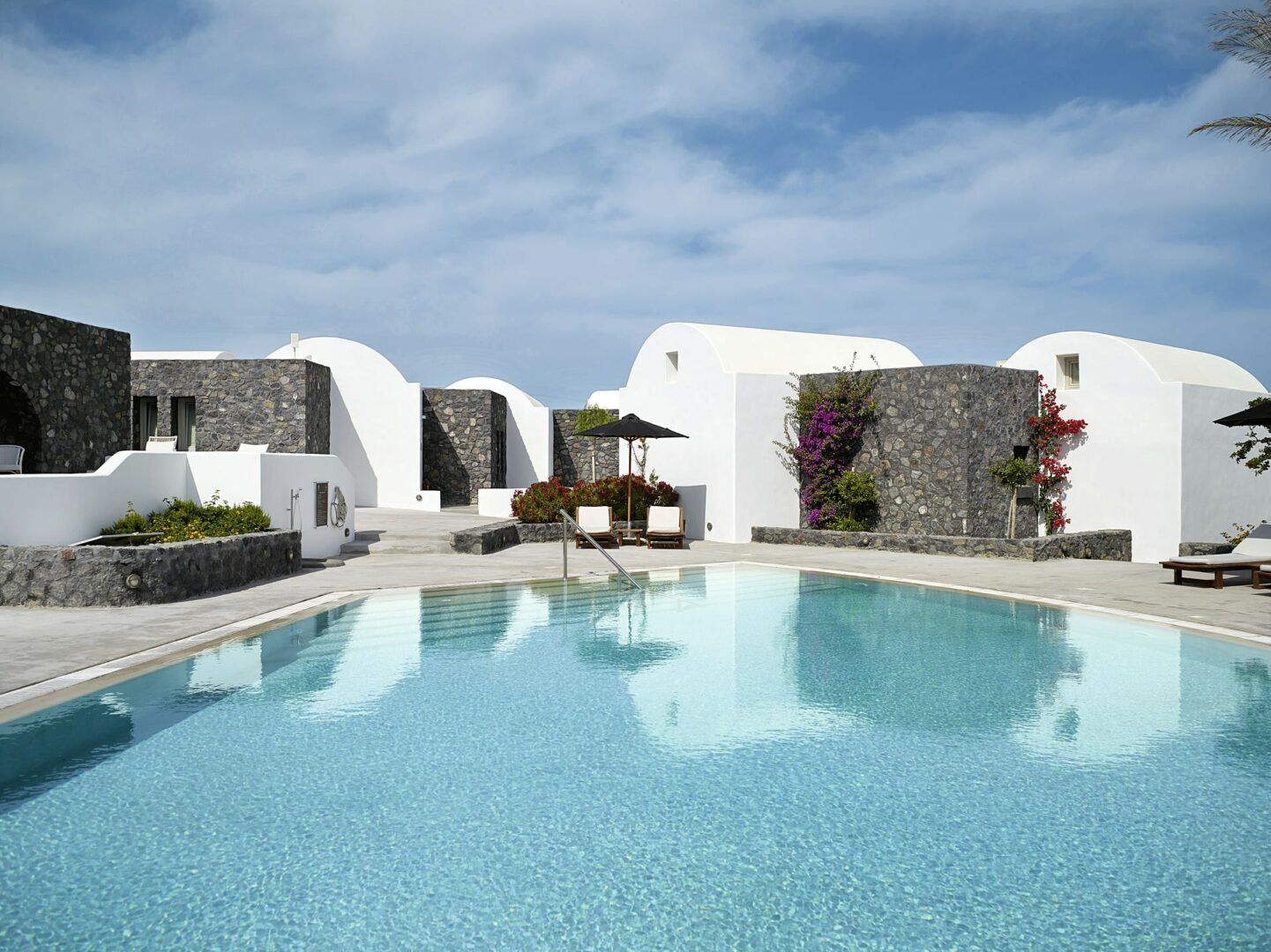 A turquoise swimming pool in front of white, domed buildings with dark stone walls under a blue sky. Andronis Luxury Suites