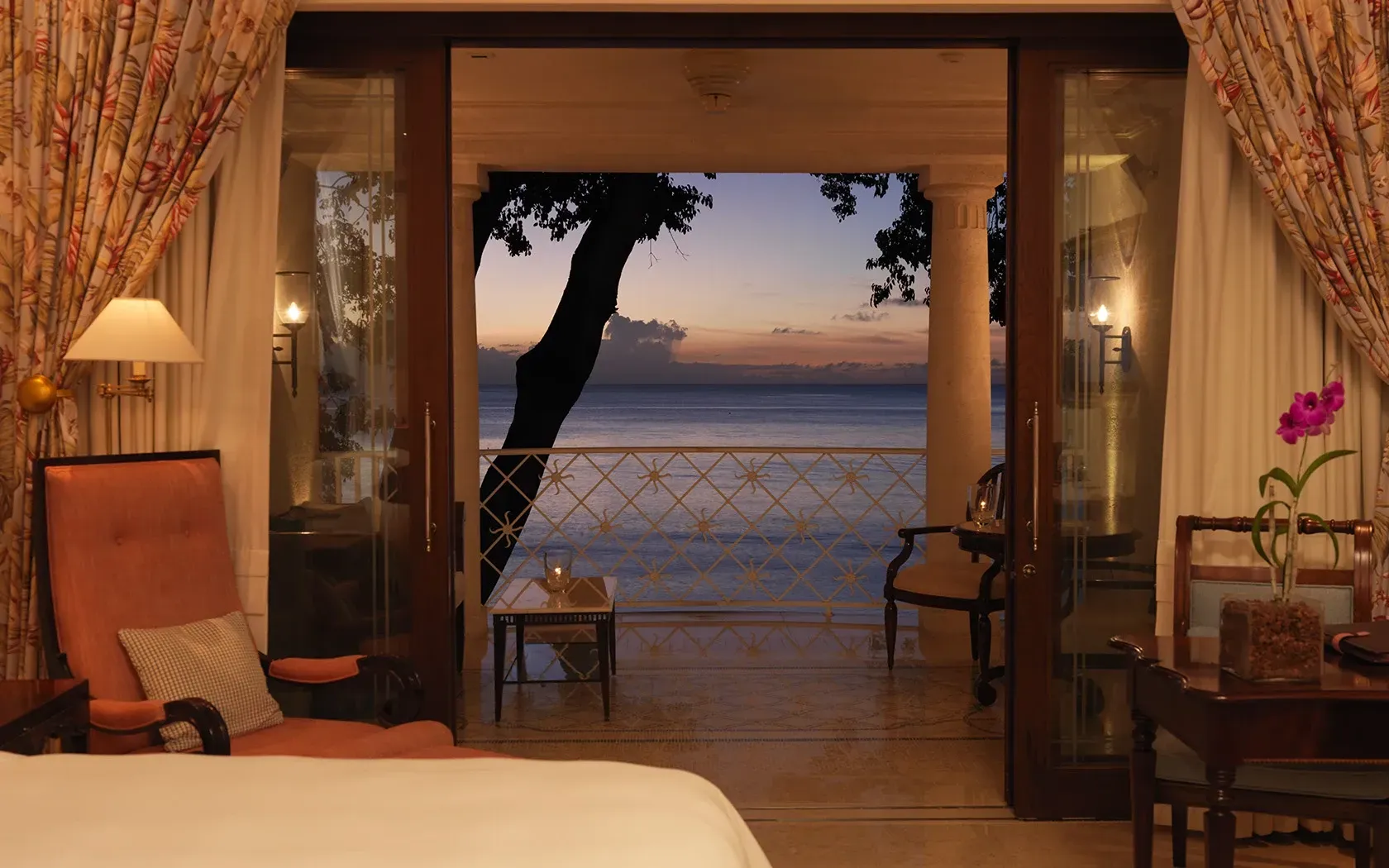 A hotel room view through open glass doors to a balcony overlooking the ocean at sunset with a tree in silhouette.