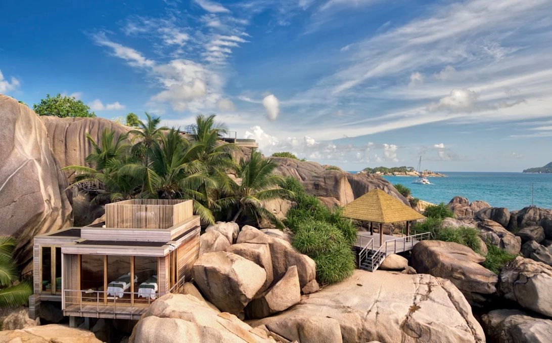 A modern resort villa with wooden accents nestled among large boulders, lush palm trees, and a tropical ocean view.