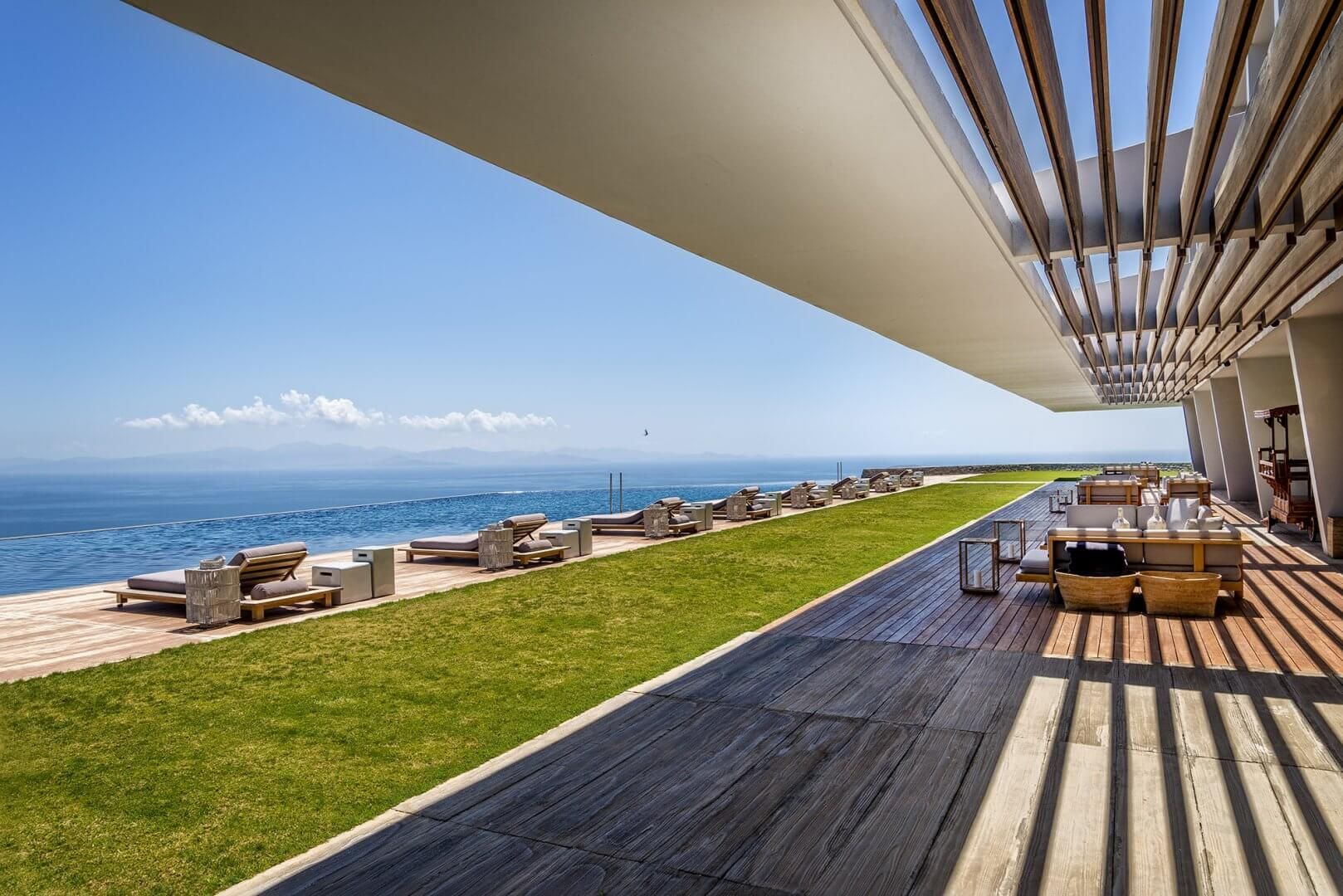 A modern terrace with outdoor furniture overlooking the ocean, featuring a grassy lawn and a slatted wooden overhang.