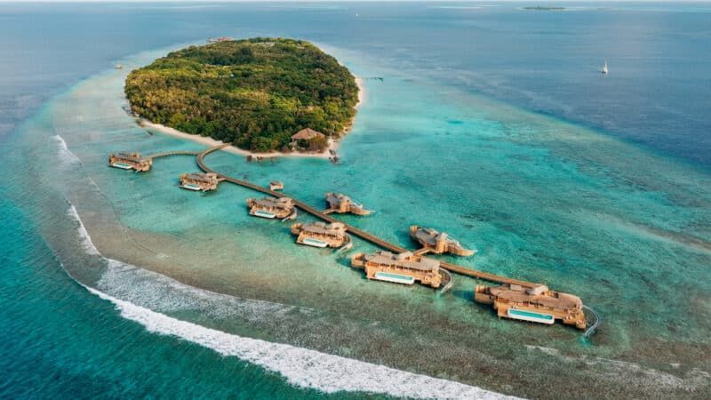 Overwater bungalows with thatched roofs in turquoise water, island in the background, blue sky. Soneva Fushi