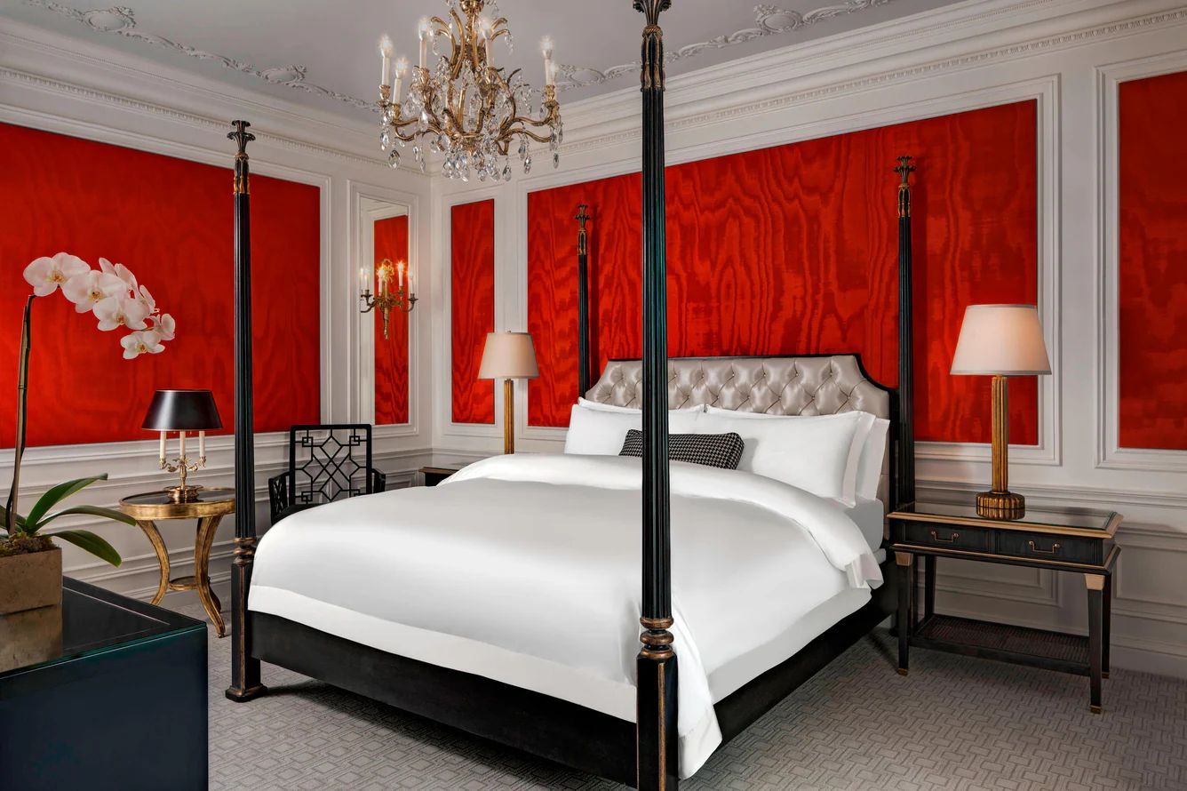 Luxurious bedroom with four-poster bed, red walls, chandelier, and side tables with lamps. St. Regis New York hotel