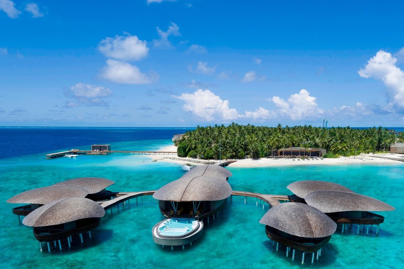 Overwater bungalows with thatched roofs in turquoise water, island in the background. Sunny day. Maldives