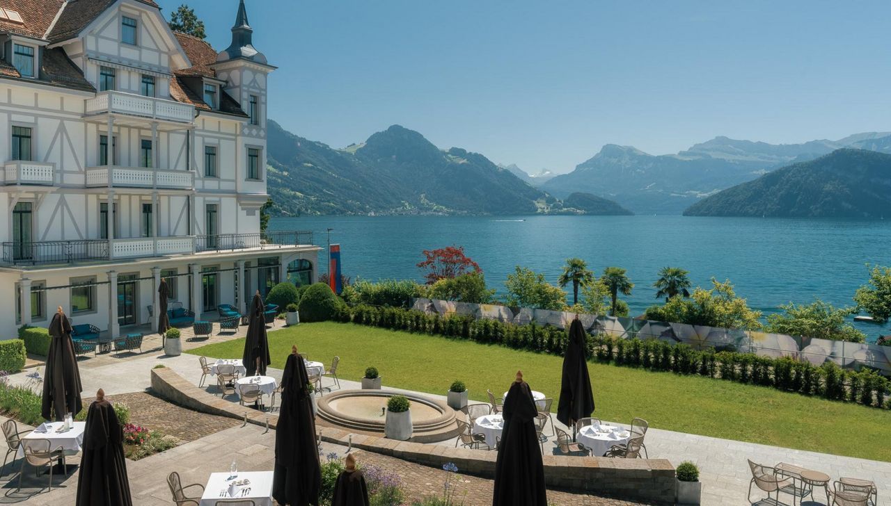 Hotel with outdoor seating overlooking a lake and mountains on a sunny day.