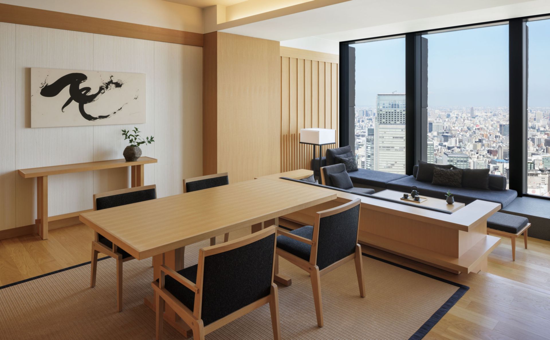 A bright, minimalist hotel suite featuring a wooden dining set, a window-side lounge area, and a city view.