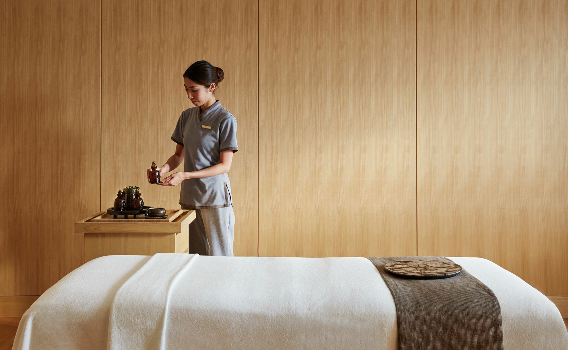 Woman preparing massage table in a spa. Wooden walls, beige linens, and dark accessories are visible. Aman Tokyo
