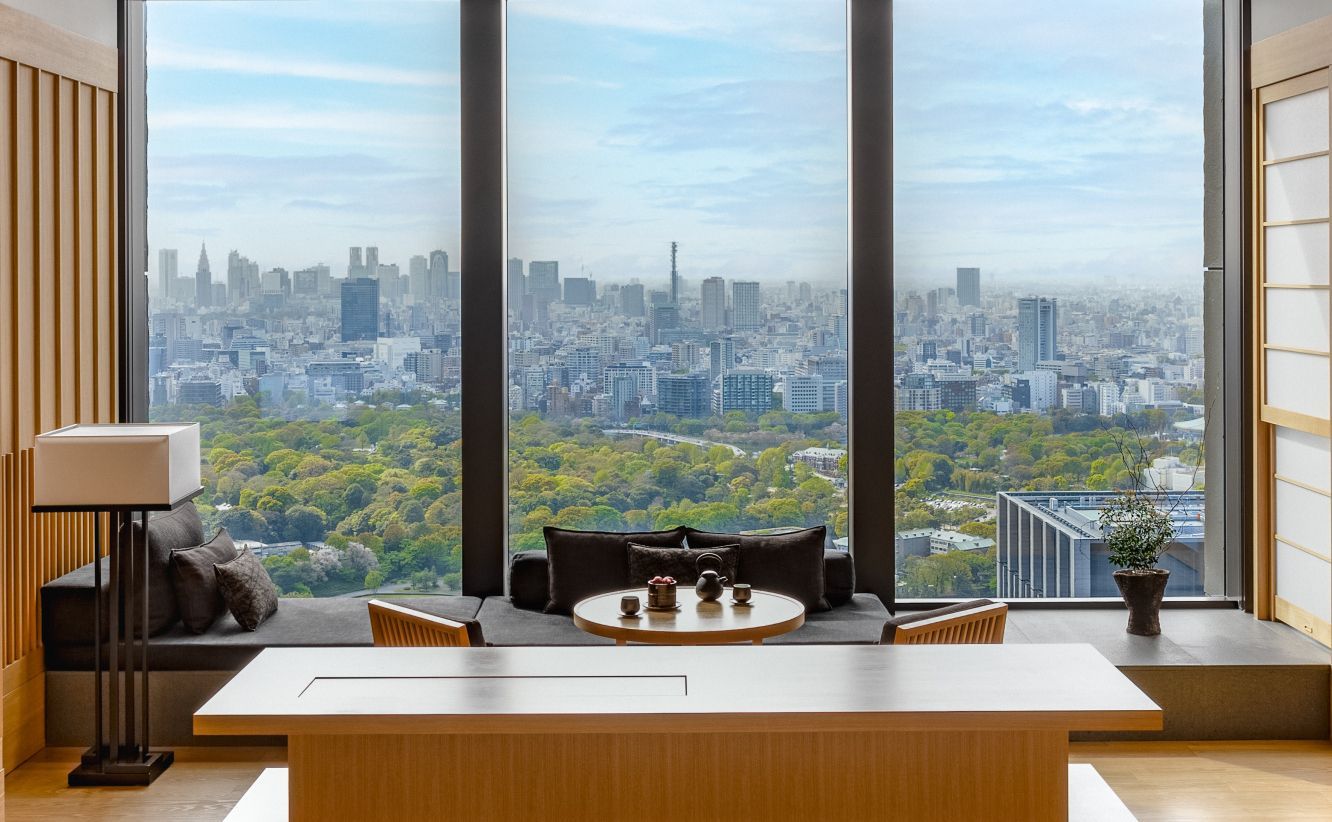 A modern room with a cityscape view through a large window; a wooden table, seating area, and lamp are visible. Aman Tokyo