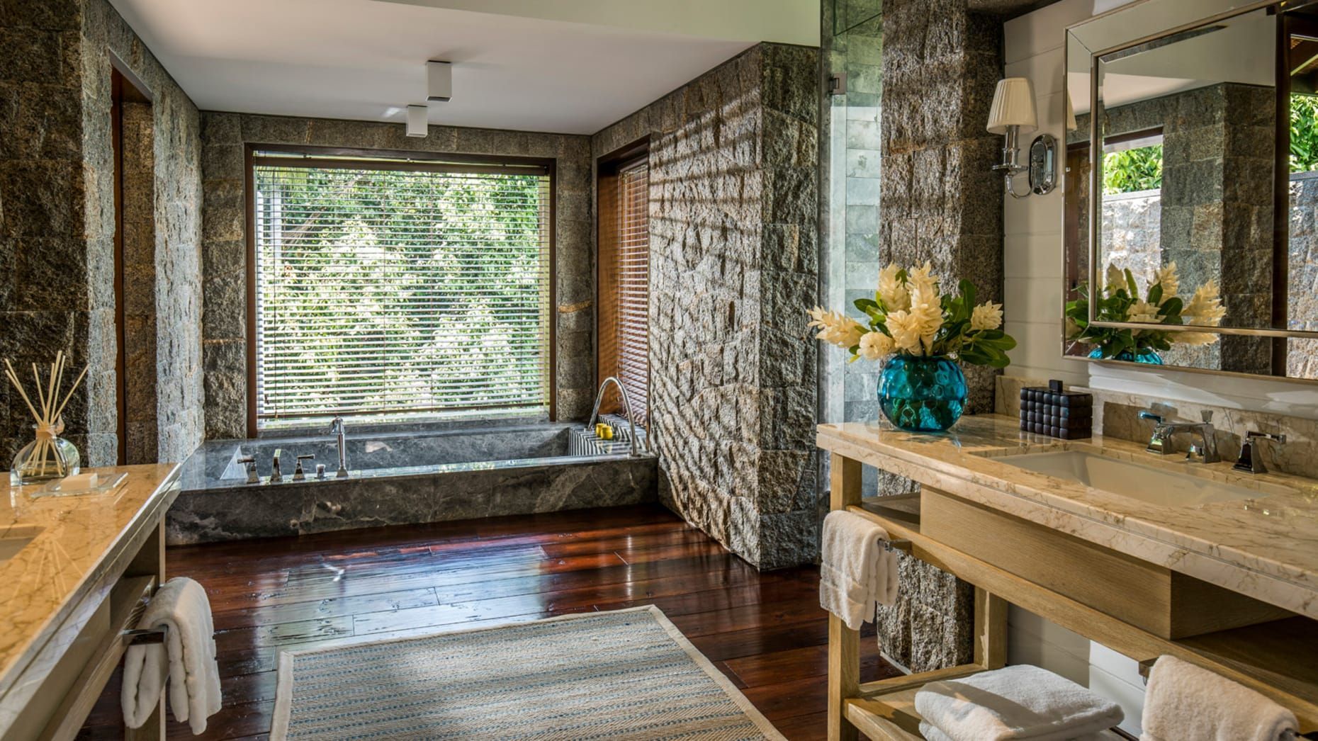 Luxury bathroom with stone walls, wooden floor, large window overlooking greenery, and a stone bathtub.