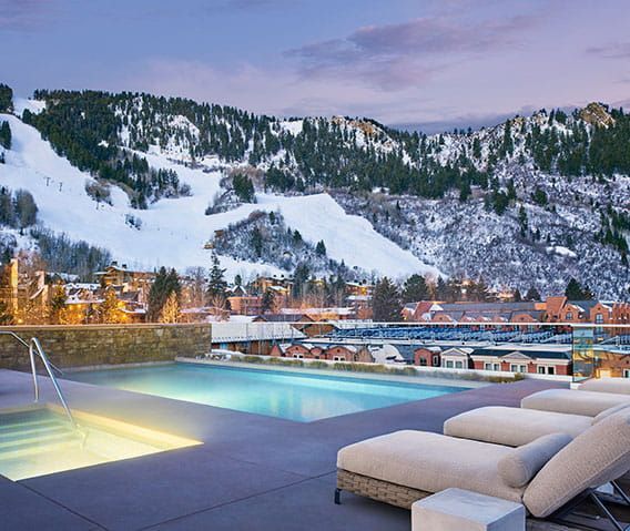 Rooftop pool and hot tub overlooking snow-covered mountains and town at dusk. Lounge chairs are visible. Aspen Snowmass, Colorado