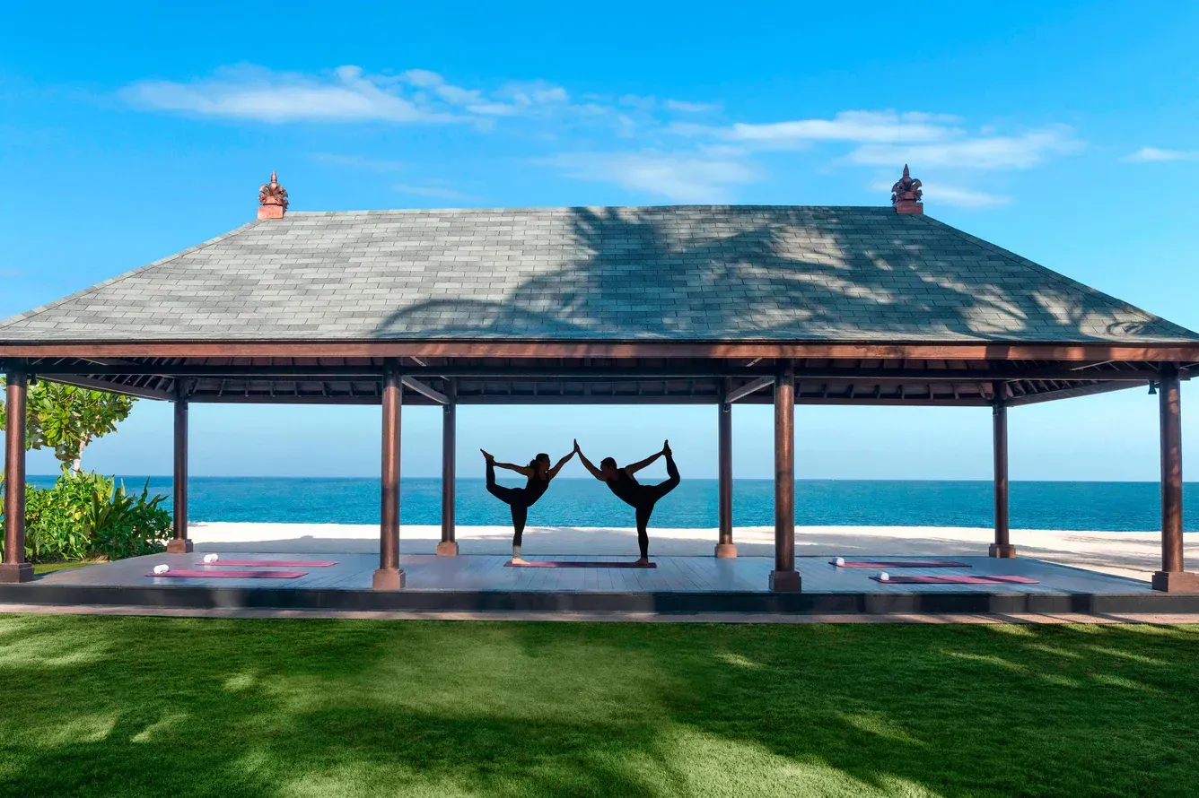 Two silhouetted dancers pose under a beach pavilion with ocean and blue sky beyond.