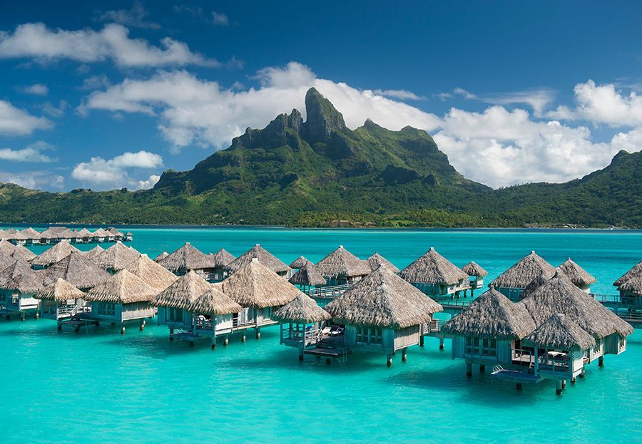 Overwater bungalows in turquoise water with lush green mountain backdrop under a blue sky. Bora Bora