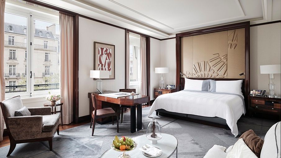 Luxurious hotel bedroom with bed, desk, seating area, and large windows overlooking a city. The Peninsula Paris