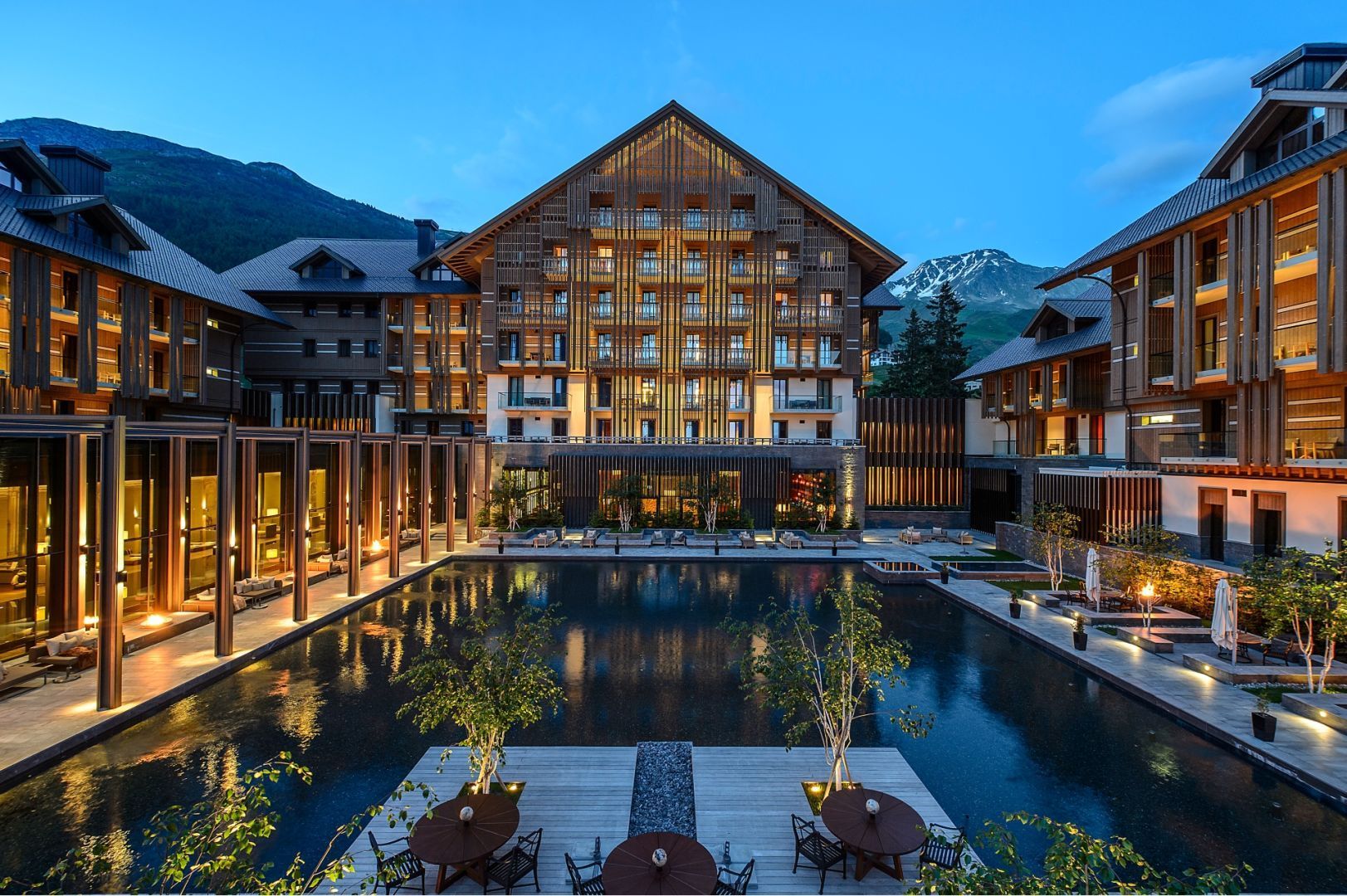 Luxury hotel with wooden exterior, pool, and mountain backdrop at dusk.  The Chedi Andermatt_