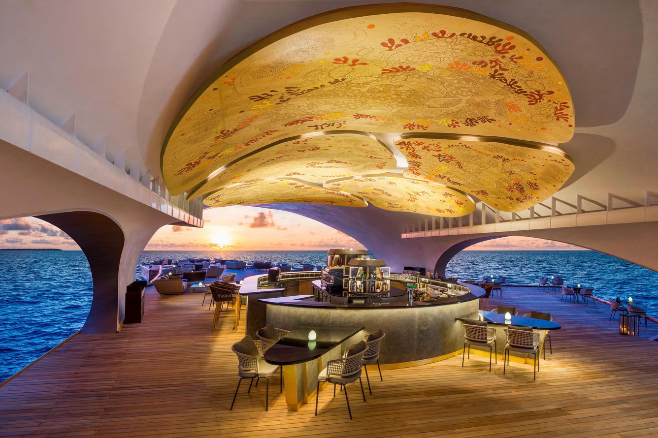 Coastal bar with sunset view, featuring a curved ceiling adorned with textured panels, a circular bar, and outdoor seating.