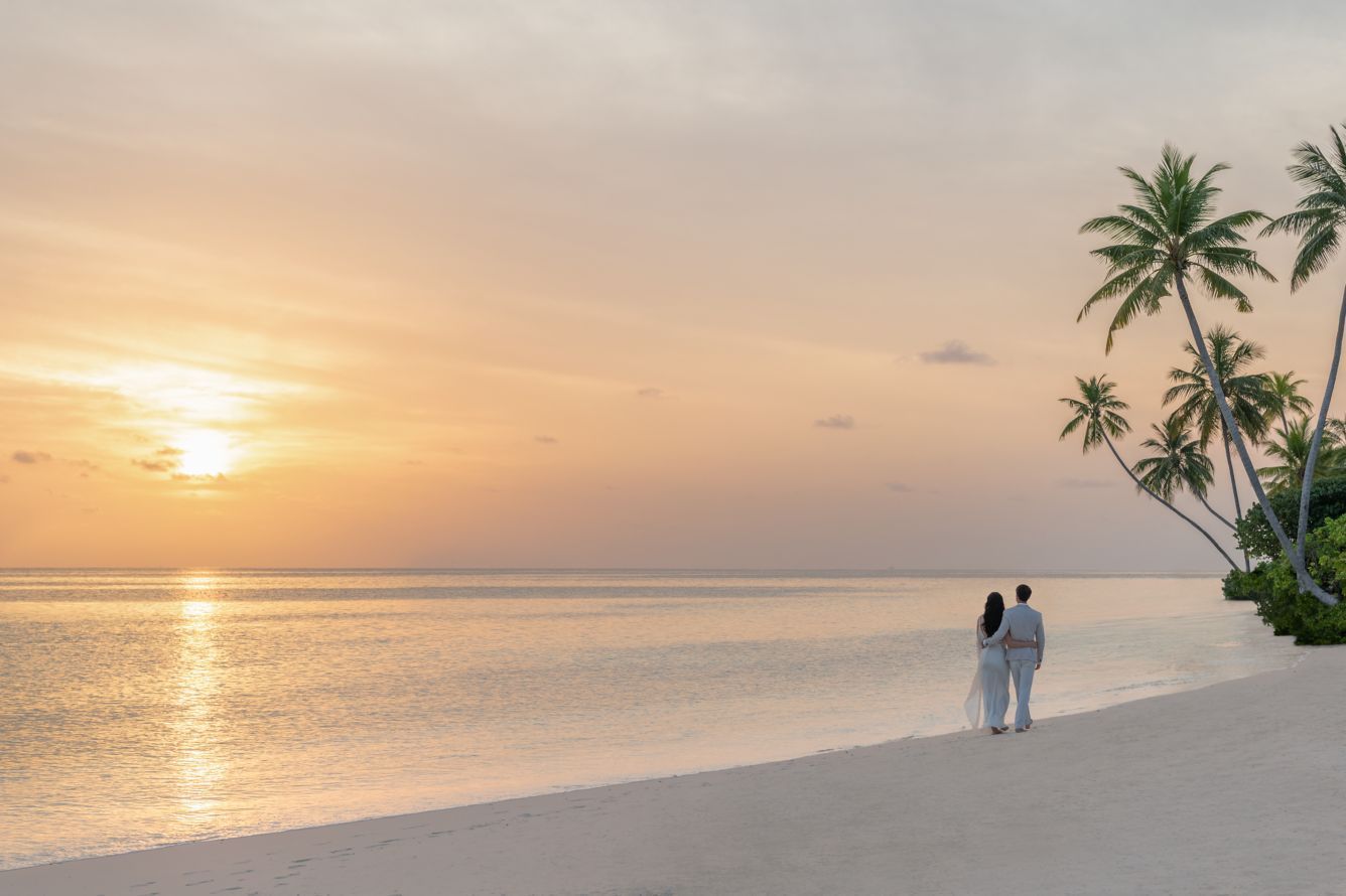 Couple standing on a white sand beach, watching sunset over the calm ocean, with palm trees.