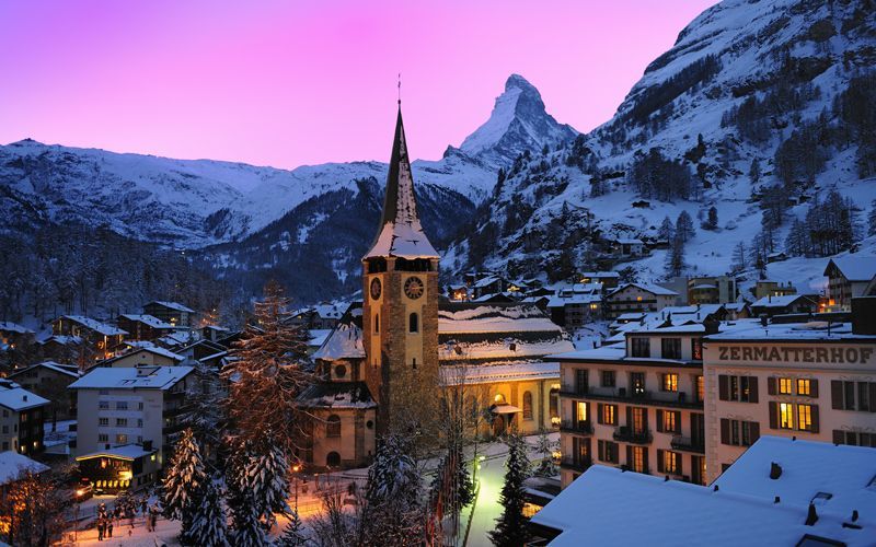 Snowy village nestled against a mountain, with a church steeple and pink-hued twilight sky. Zermatt