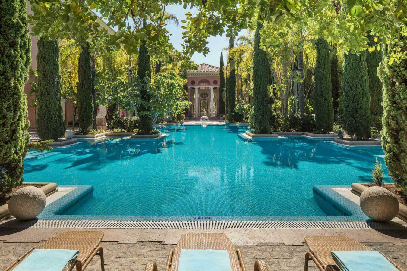 A turquoise pool surrounded by tall trees and a building in the background; two lounge chairs in the foreground.