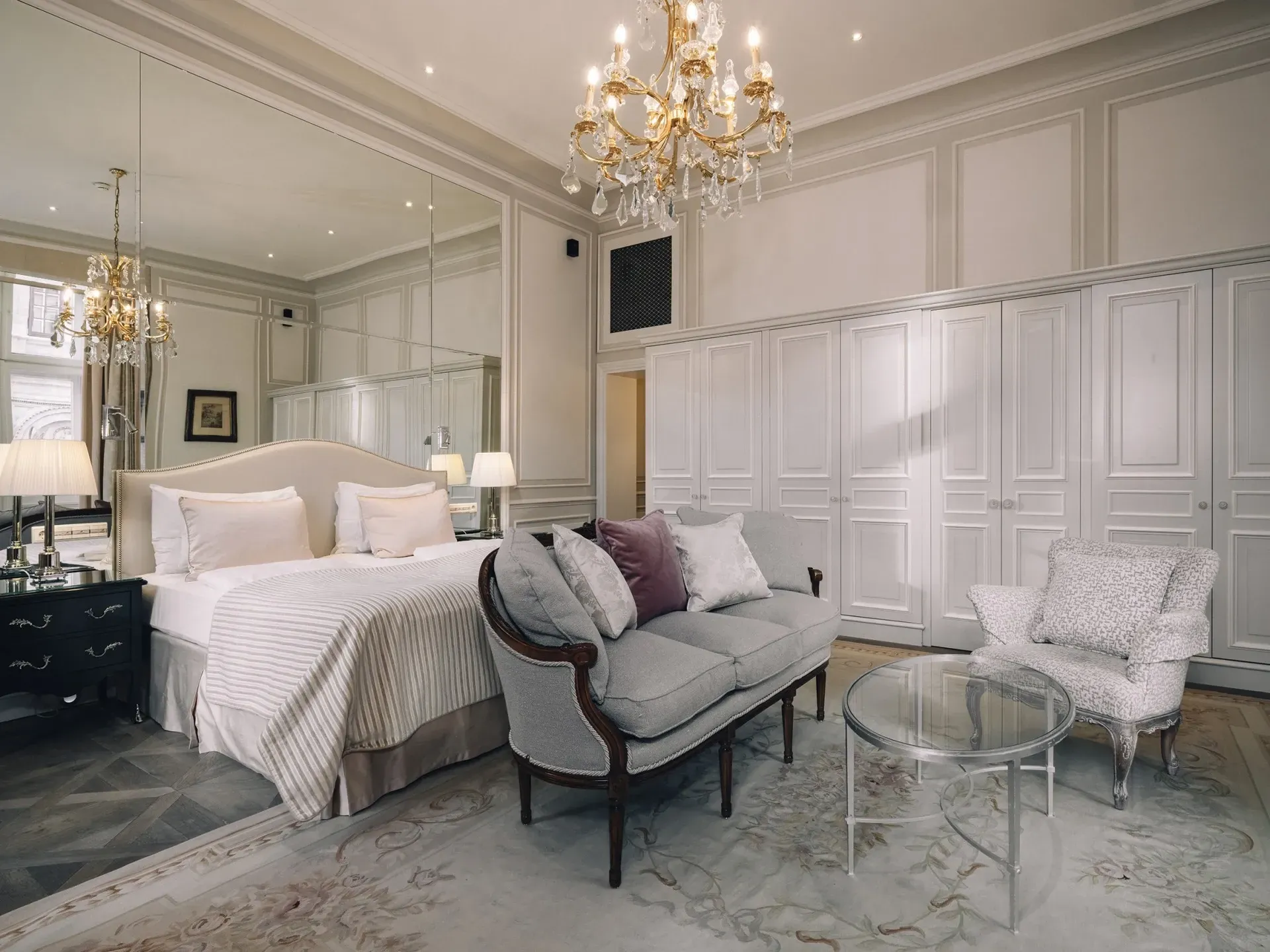 Elegant bedroom with white paneled walls, chandelier, tufted bed, and mirrored seating area