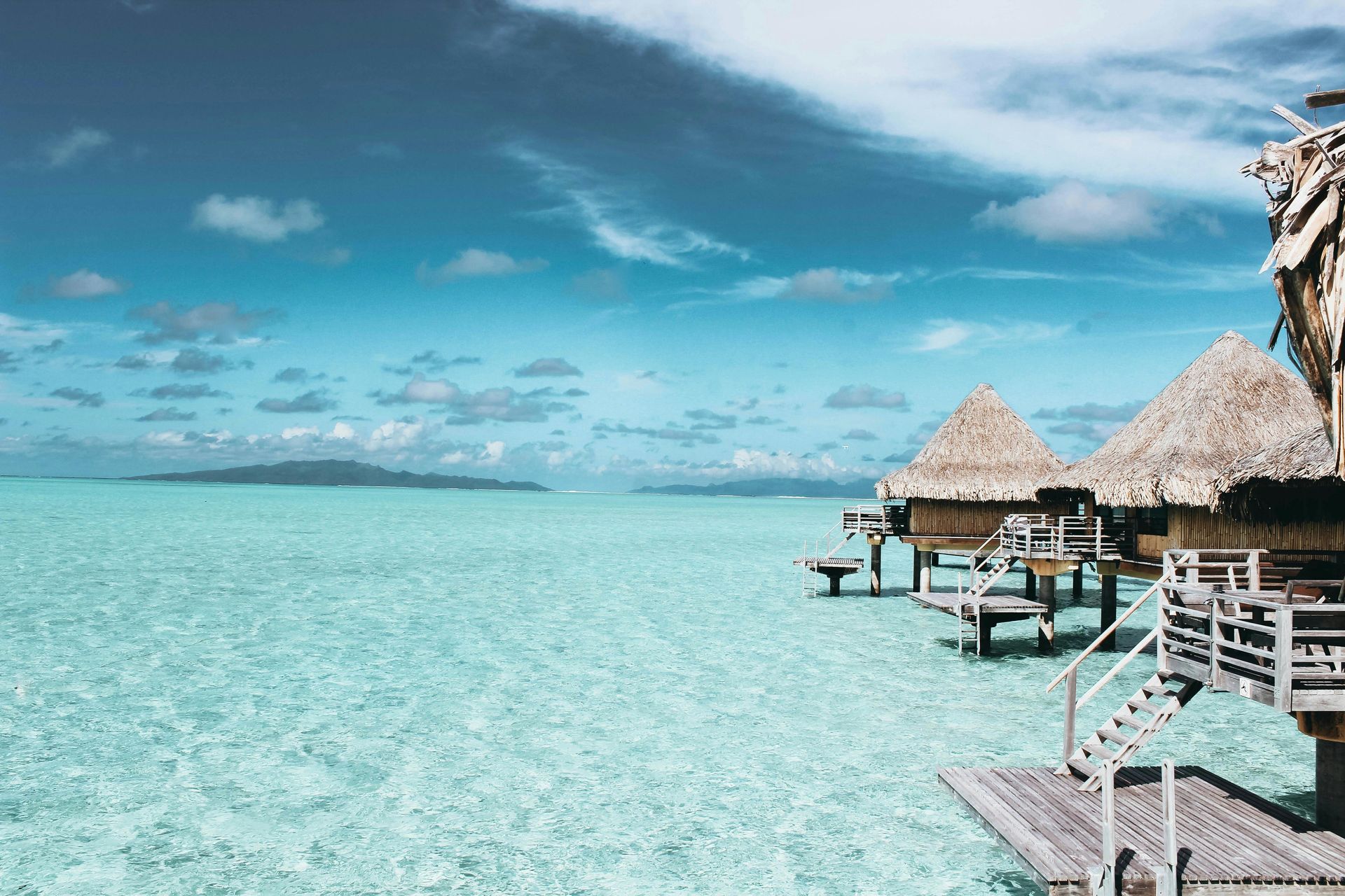 Overwater bungalows with thatched roofs in turquoise water under a partly cloudy sky. Bora Bora