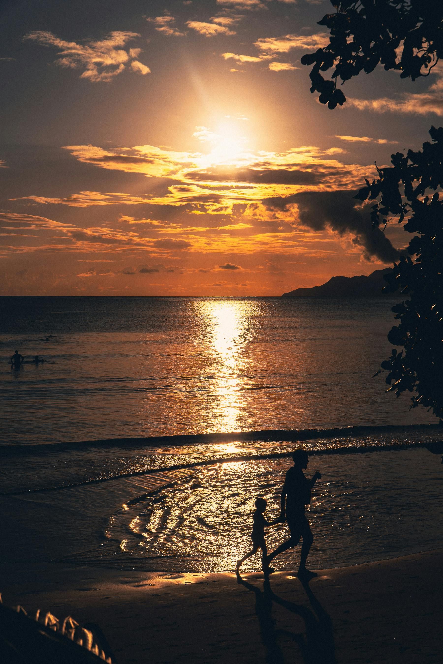 Sunset over ocean; silhouette of person holding a child on a beach.