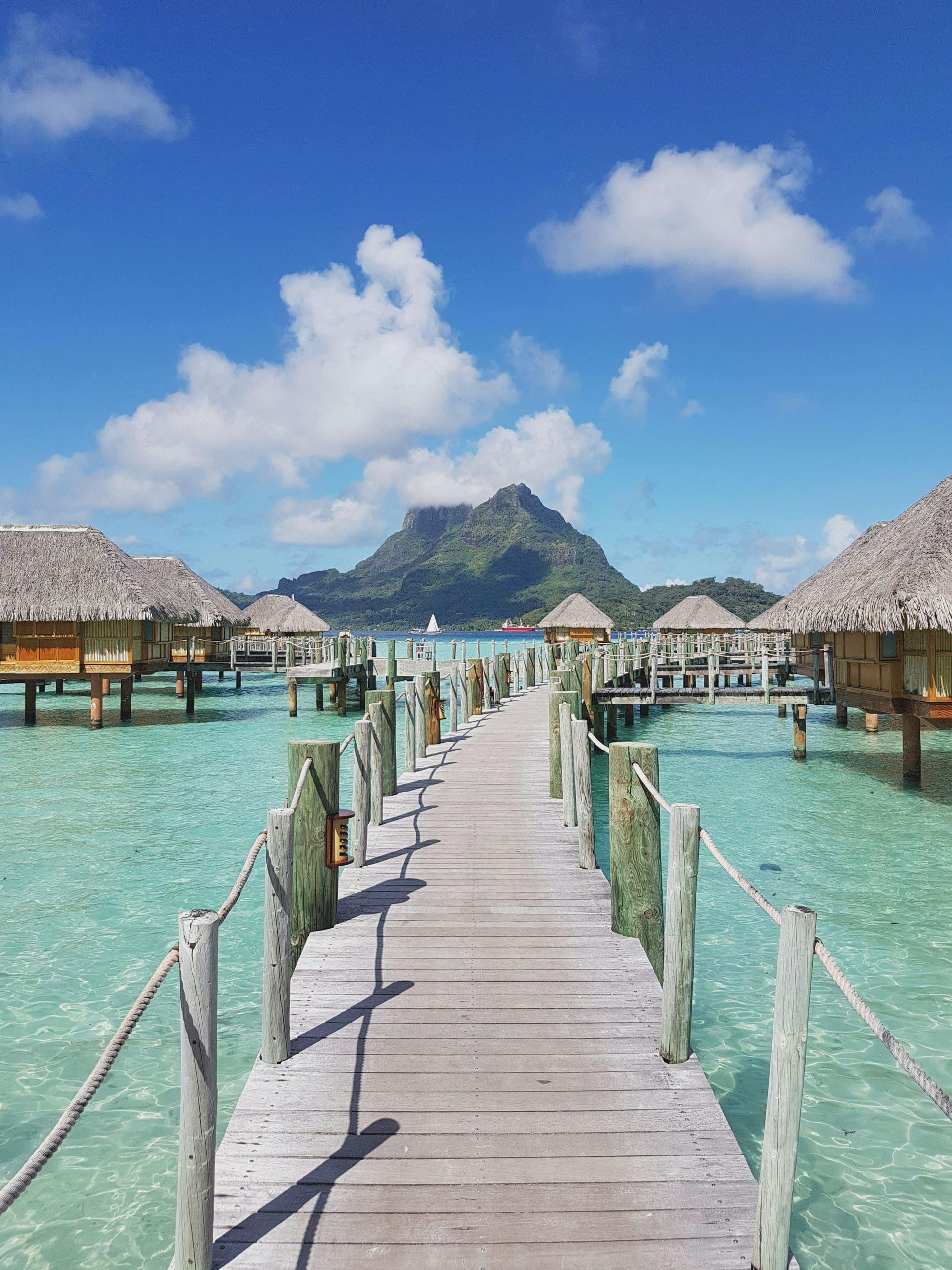 Wooden pier leading to overwater bungalows in turquoise water; mountain in the background under a blue sky.