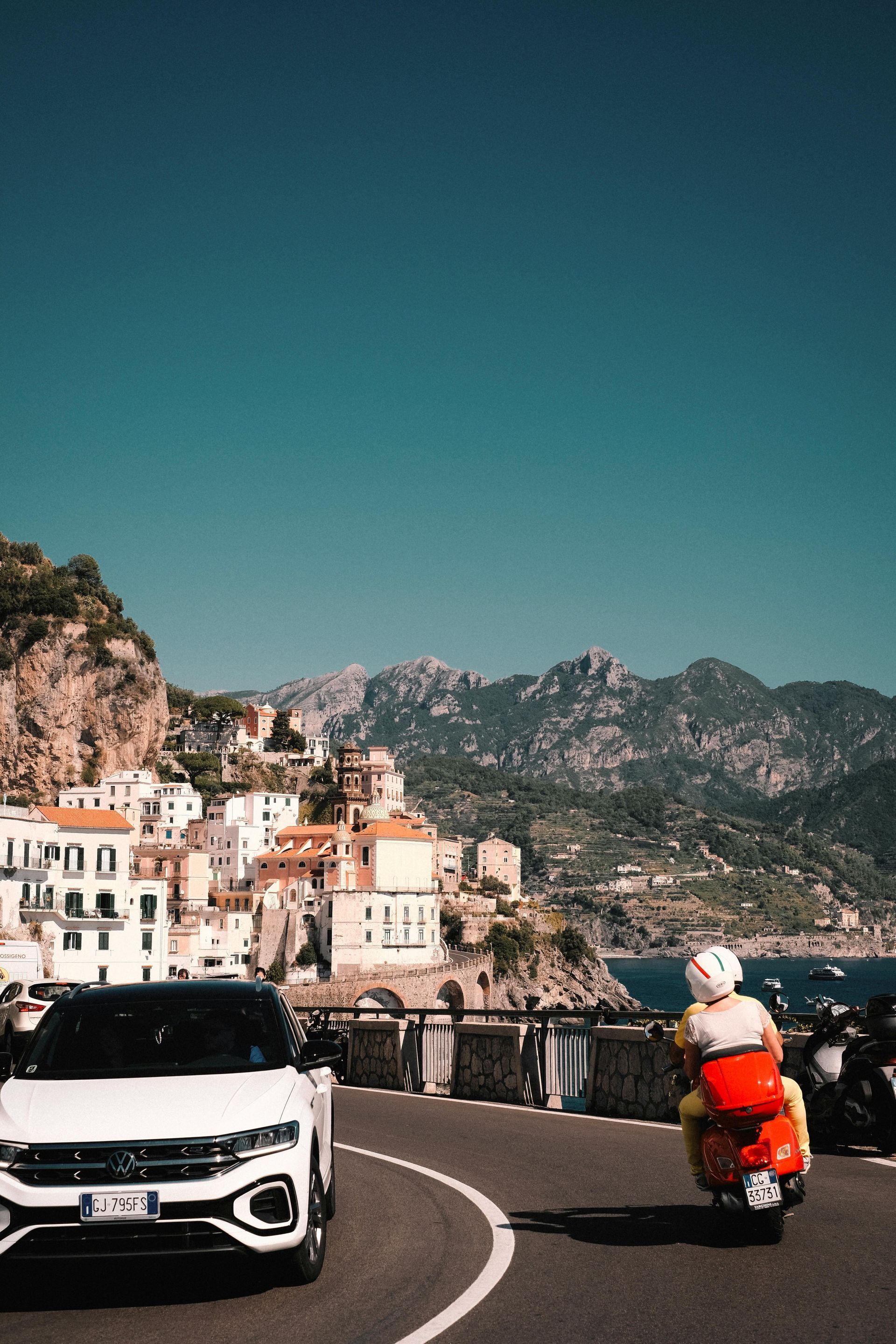 White car and red scooter on a coastal road with Italian buildings, mountains, and blue sky.
