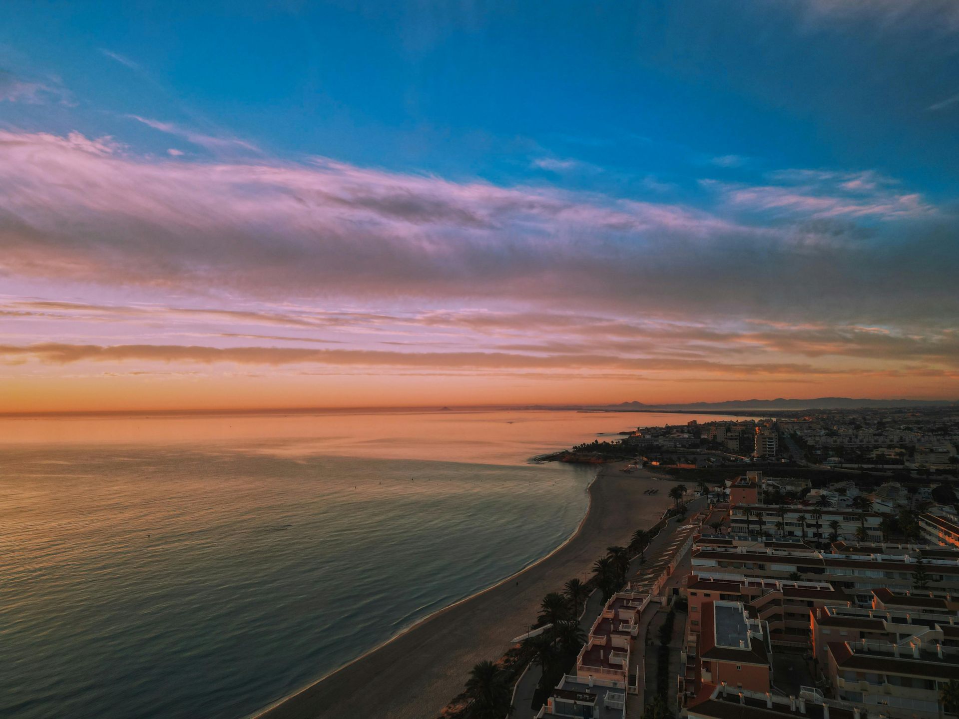 Aerial view of a sunset over a calm ocean, sandy beach, and coastal town with warm orange and purple clouds.