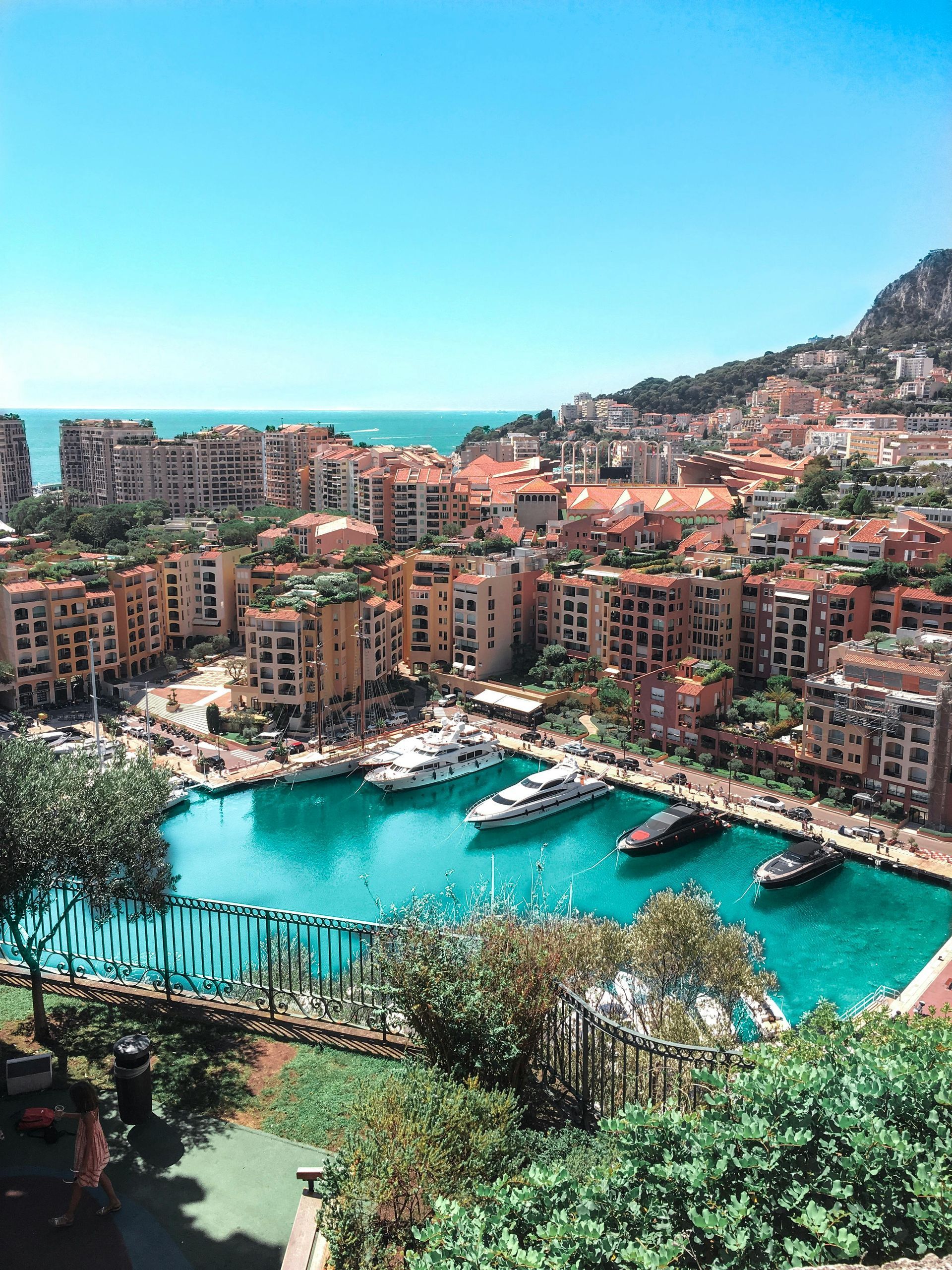 Coastal cityscape with buildings overlooking a turquoise harbor filled with boats under a blue sky.