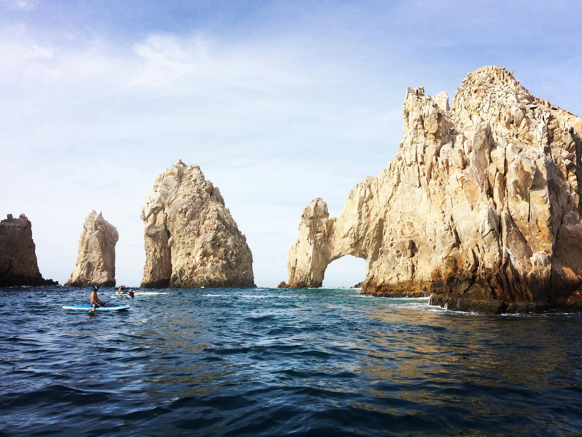 Rocky sea arches and cliffs rising from a blue ocean under a clear sky, with a small kayak in the water