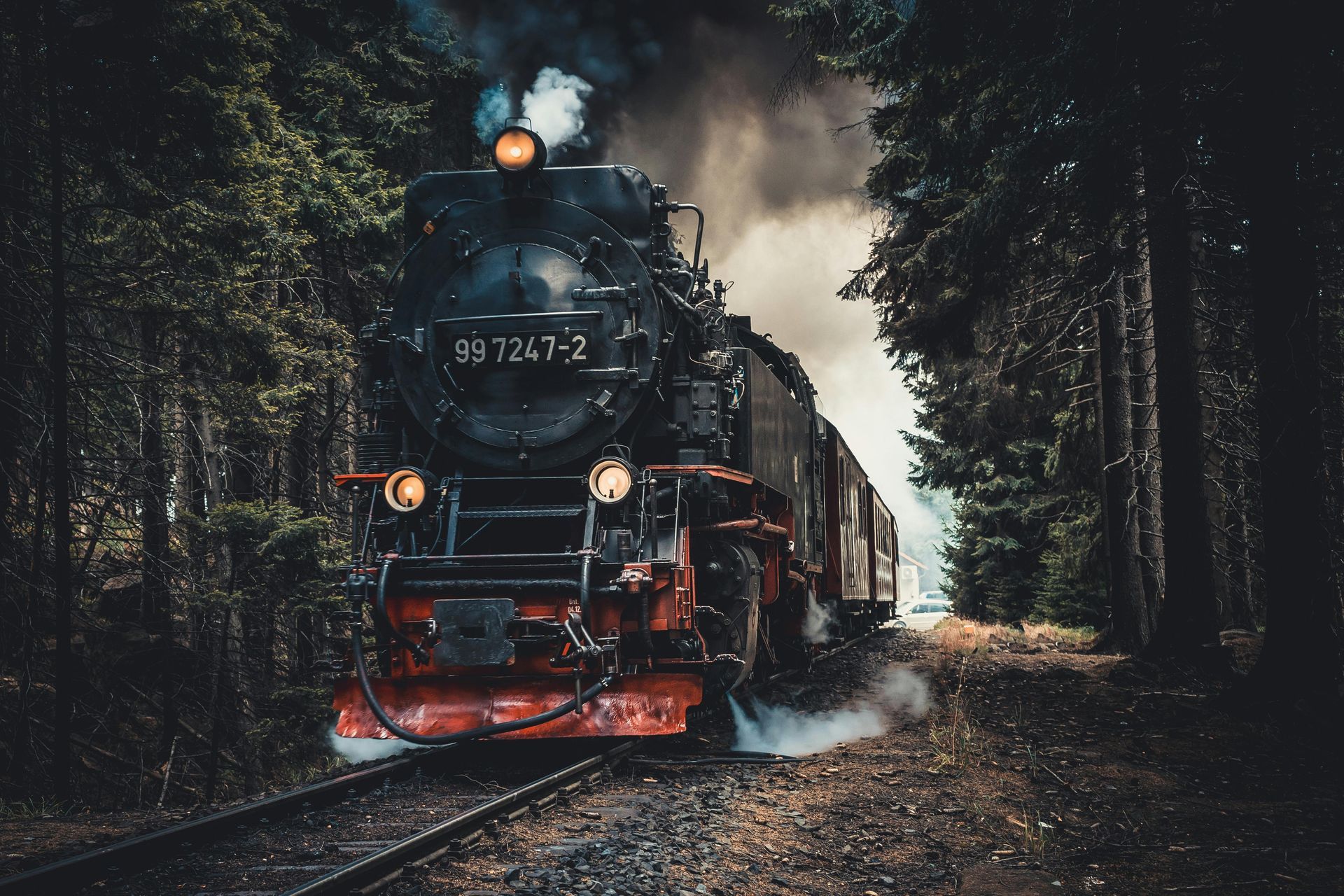 A black steam locomotive travels through a dark, foggy forest, with smoke billowing from its stack and headlamps glowing.