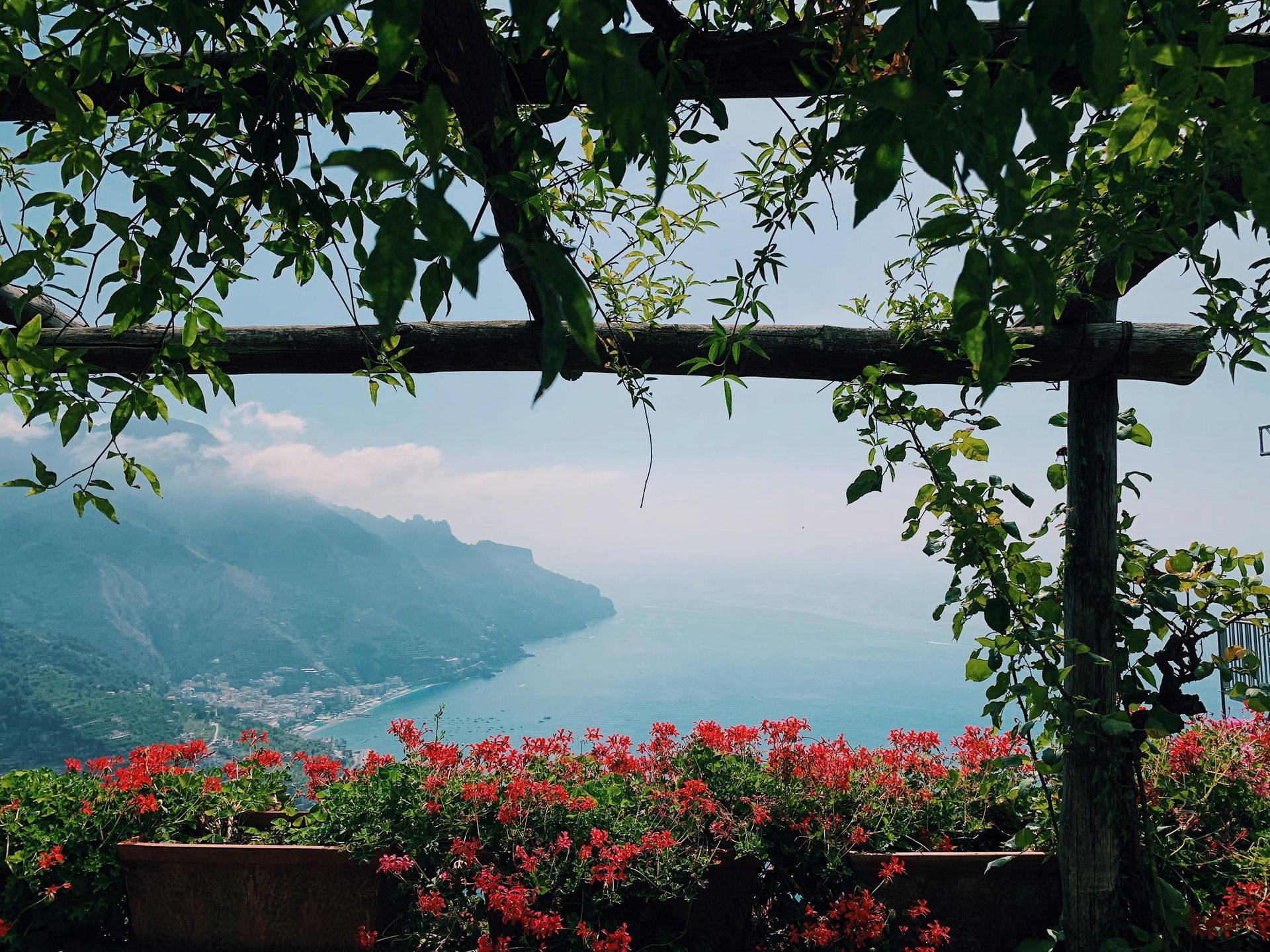 Vine-covered pergola framing a coastal view. Red flowers in foreground, blue sea and mountains in the distance. Ravello: Hilltop Elegance