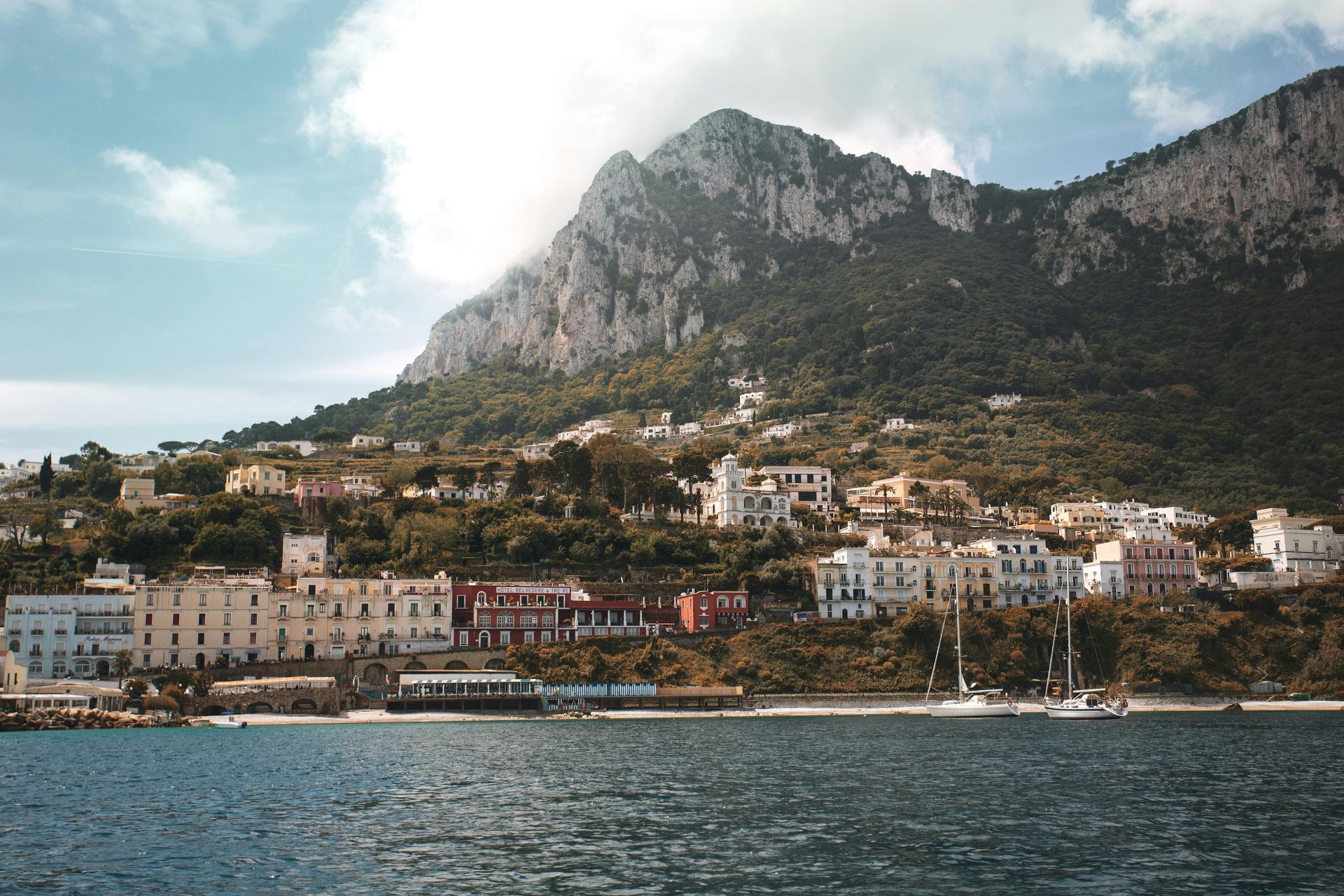 Coastal town nestled at the base of a large mountain, with blue water and scattered clouds. Capri