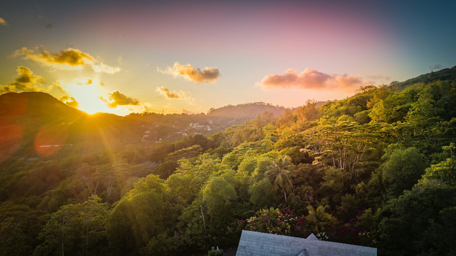 Sunset over lush, green hillside with a building in the foreground; sunbeams burst through the clouds.