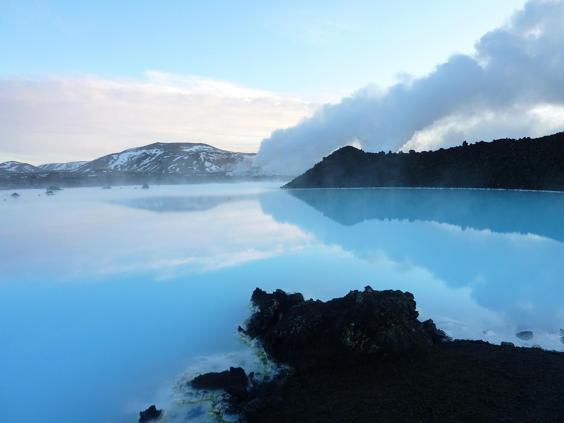 Blue lagoon with steaming water and dark volcanic rock. Snow-covered hills in the distance under a blue sky.